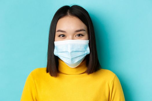 Covid-19, social distancing and pandemic concept. Close up of young asian woman with short dark hair, wearing medical mask and smiling with eyes, looking hopeful at camera.