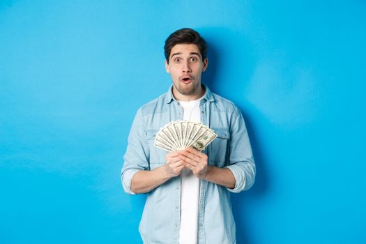 Concept of finance, credit and banking. Surprised man holding money, looking at camera wondered, standing over blue background.