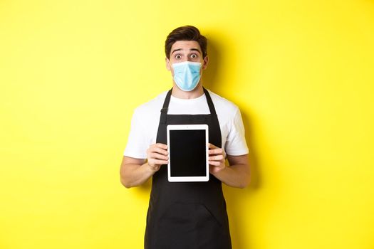 Concept of covid-19, small business and pandemic. Surprised waiter in black apron and medical mask showing tablet screen, standing over yellow background.