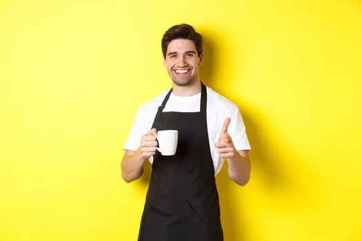 Barista bringing coffee and pointing finger gun at camera, standing in black apron against yellow background.
