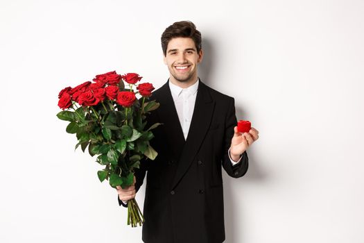 Image of handsome man in black suit, holding bouquet of red roses and a ring, making a proposal, smiling confident, standing against white background.