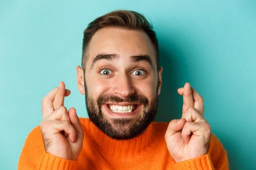 Headshot of hopeful bearded man making a wish, smiling and holding fingers crossed for good luck, standing over light blue background.