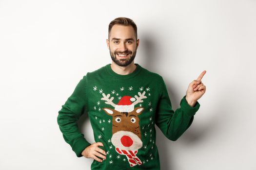 Winter holidays and christmas. Handsome bearded adult man pointing finger right at promo banner, standing in funny sweater against white background.