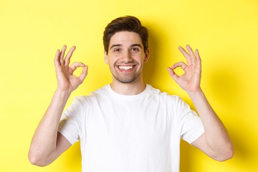 Close-up of handsome young man showing okay sign, approve and agree, smiling satisfied, standing over yellow background.