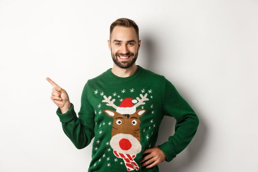 New year celebration and winter holidays concept. Confident and happy man with beard, wearing christmas sweater, pointing at upper left corner banner, white background.