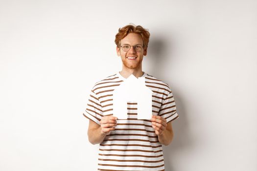 Real estate concept. Smiling redhead man in glasses holding cutout paper house and looking at camera, saving for buying property, standing over white background.