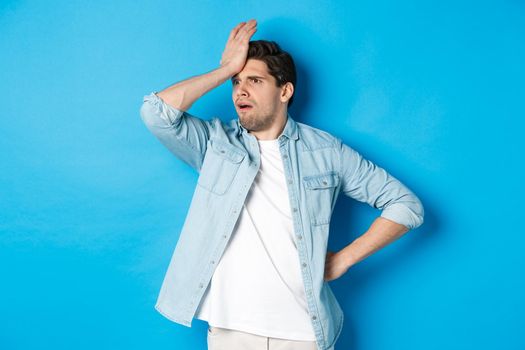 Upset man making facepalm and looking away concerned, forgot something important, slap forehead troubled, standing against blue background.