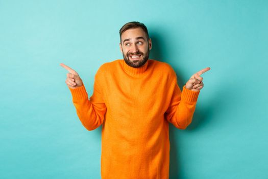 Confused handsome man shrugging indecisive, pointing fingers sideways, cant choose between two variants, standing against light blue background.