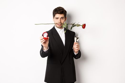 Passionate young man in suit making a proposal, holding rose in teeth and glass of champagne, showing engagement ring, asking to marry him, standing against white background.