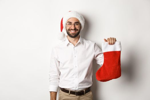 Merry christmas, holidays concept. Happy adult man receive gifts in xmas sock, looking excited, wearing santa hat, white background.