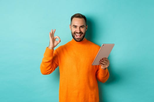 Satisfied adult man smiling, using digital tablet and showing okay sign, approve and agree, standing against turquoise background.