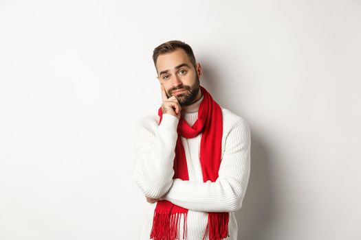 Bored young man looking indifferent and tired at camera, listening someone, standing in winter sweater and scarf, white background.