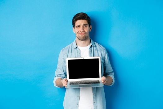 Skeptical and displeased bearded guy showing laptop screen and grimacing, having doubts, standing over blue background in casual clothes.