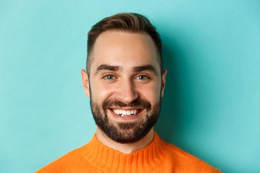 Headshot of handsome caucasian man with beard smiling happy at camera, standing in orange sweater against turquoise background.