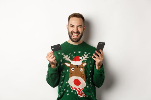 New Year, online shopping and christmas concept. Excited man using credit card and smartphone, standing over white background.