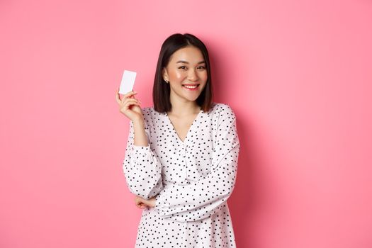 Shopping concept. Confident and happy asian woman holding credit card and smiling satisfied, standing against pink background.