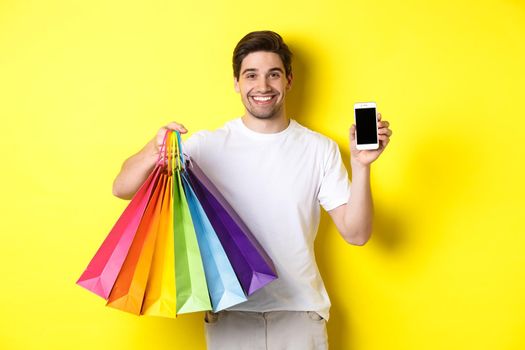 Young man holding shopping bags and showing mobile phone screen, money application, standing over yellow background.