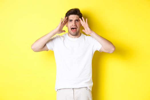 Frustrated guy looking alarmed, holding hands on head and feeling confused, panicking, standing over yellow background.