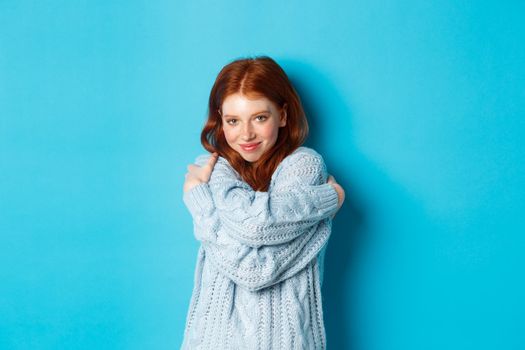 Happy cute redhead girl hugging herself, wearing comfortable and warm sweater, smiling at camera, standing over blue background.