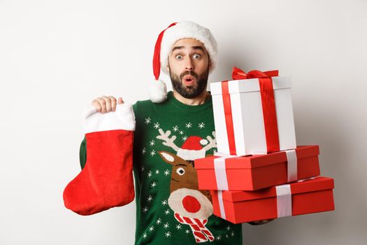 Xmas and winter holidays concept. Excited man holding christmas sock and gift boxes, celebrating New Year, bringing presents under tree, standing over white background.