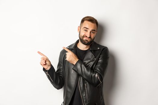 Skeptical and disapleased man in leather jacket, pointing fingers at upper left corner, showing bad promo offer, standing over white background reluctant.