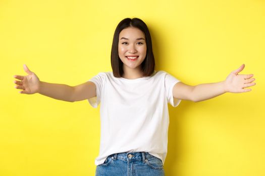 Beauty and fashion concept. Friendly asian woman spread out hands and smiling, waiting for hugs, inviting you, welcome someone and looking happy, standing over yellow background.