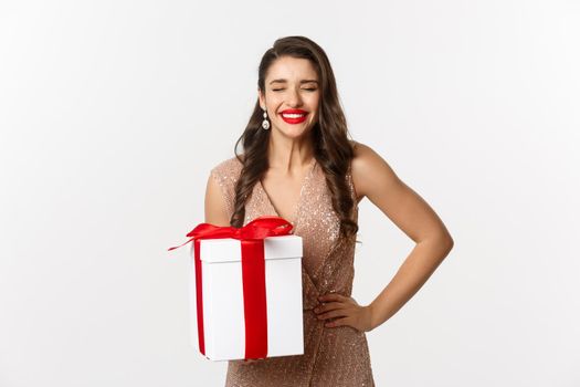 Merry Christmas. Image of attractive woman in luxury dress, celebrating winter holidays, holding gift and laughing from happiness, standing over white background.