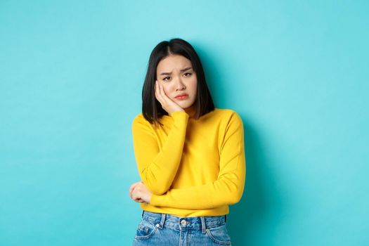 Sad and bored asian girl looking reluctant and unamused at camera, leaning face on hand, standing over blue background.