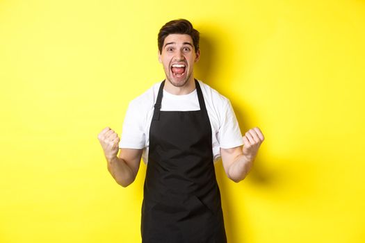 Excited coffee shop owner in black apron celebrating, making fist pump and shouting for joy, achieve goal, standing against yellow background.