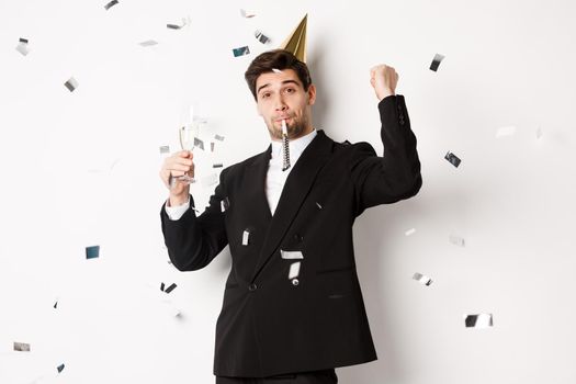 Handsome party guy in black suit having fun, celebrating new year, blowing whistle and drinking champagne while confetti falling, standing happy against white background.