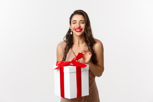 Merry Christmas. Charming woman in elegant dress open gift box and smiling delighted, unwrapping a present, standing over white background.