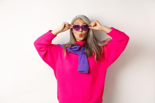 Fashionable asian senior woman in sunglasses and stylish pink sweater pucker lips, raising eyebrows and looking surprised at camera, white background.