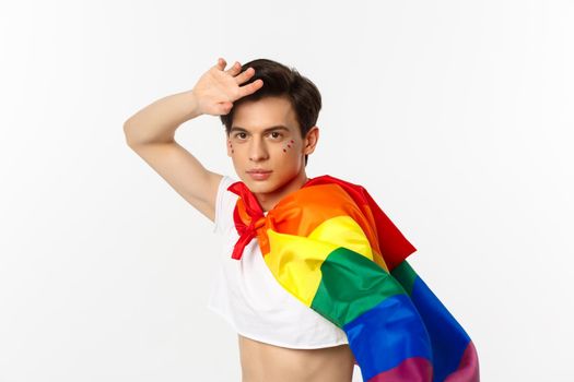 Beautiful gay man with glitter on face, wearing crop top and rainbow lgbt flag, posing against white background.