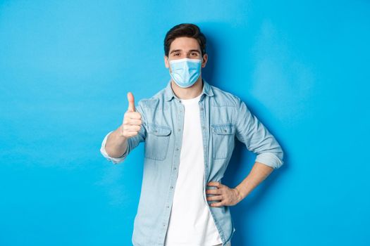 Concept of covid-19, pandemic and social distancing. Satisfied guy in medical mask showing thumb up in approval, standing against blue background.