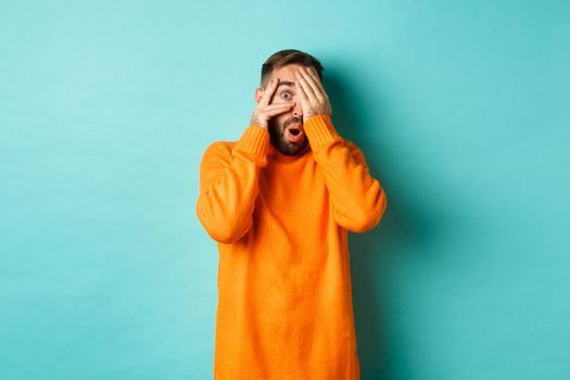 Curious man peeking at something, shut eyes but looking through fingers with amazement and interest, standing over light blue background.