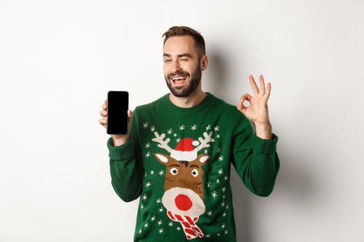 Christmas holidays and shopping concept. Confident guy winking and showing okay sign with mobile screen, standing over white background.