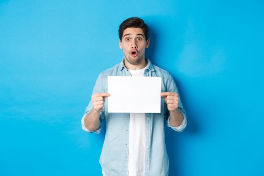 Surprised man gasping and looking impressed at camera, showing blank piece of paper for your sign, standing over blue background.