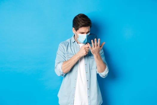 Concept of coronavirus, social distancing and pandemic. Man in medical mask looking at his palm through magnifying glass, standing over blue background.