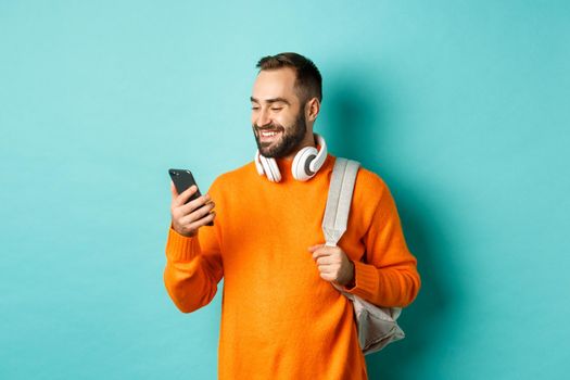 Caucasian man with headphones and backpack looking at phone, reading message and smiling, standing over turquoise background.