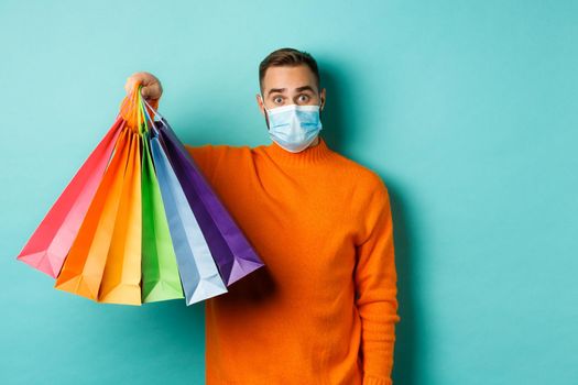 Covid-19, social distancing and lifestyle concept. Young man in face mask showing shopping bags, buying holiday gifts during pandemic, standing over blue background.