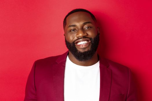 Close-up of handsome bearded Black man celebrating New Year, wearing party outfit and smiling happy, standing over red background.