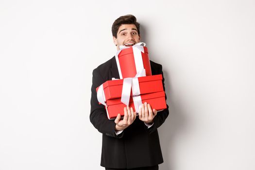 Concept of christmas holidays, celebration and lifestyle. Image of happy man in suit carry presents for new year, holding boxes with gifts and smiling, standing against white background.