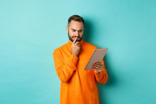 Serious man pondering and looking at digital tablet screen, reading social media, standing over turquoise background. Copy space