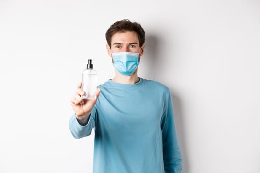 Covid-19, health and quarantine concept. Young man in medical mask stretch out hand and showing sanitizer, recommending antiseptic, standing over white background.