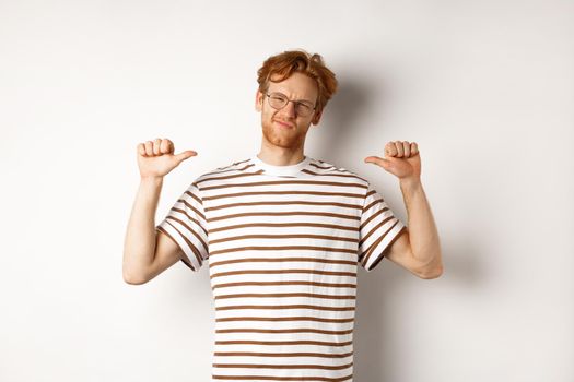 Confident young nerdy guy in glasses with red hair, pointing at himself and looking like professional, white background.