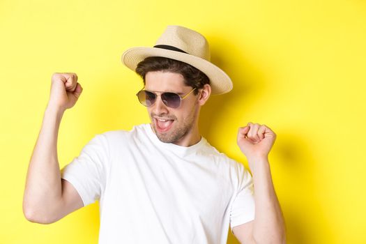 Concept of tourism and vacation. Close-up of man enjoying holidays on trip, dancing and pointing fingers sideways, wearing sunglasses with straw hat, yellow background.