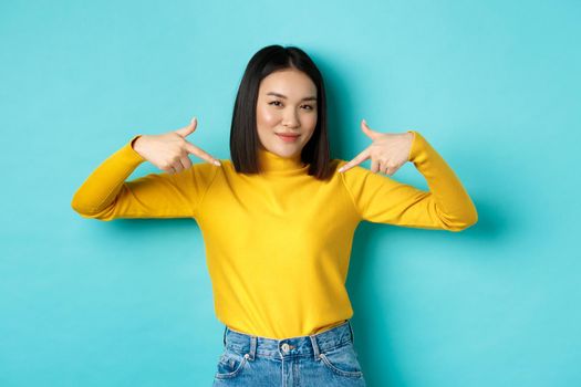 Confident asian woman in stylish sweater pointing fingers at logo on center, smiling assertive at camera, standing over blue background.