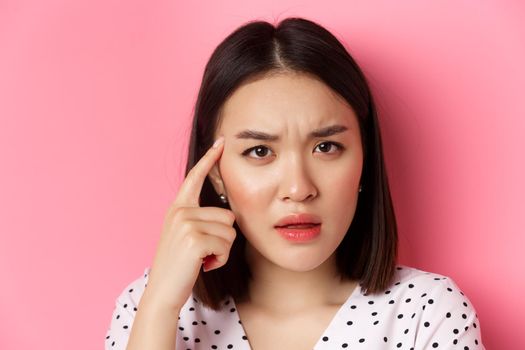 Beauty and skin care concept. Headshot of cute asian woman staring confused, pointing finger at head at frowning, standing over pink background.