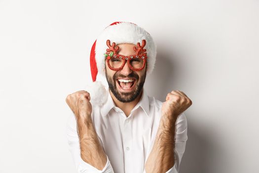 Close-up of man celebrating christmas or new year, wearing xmas party glasses and santa hat, rejoicing and shouting of joy, standing over white background.