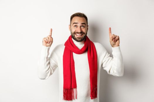 Christmas shopping and winter holidays concept. Happy bearded man in red scarf pointing fingers up, looking excited at copy space, standing over white background.
